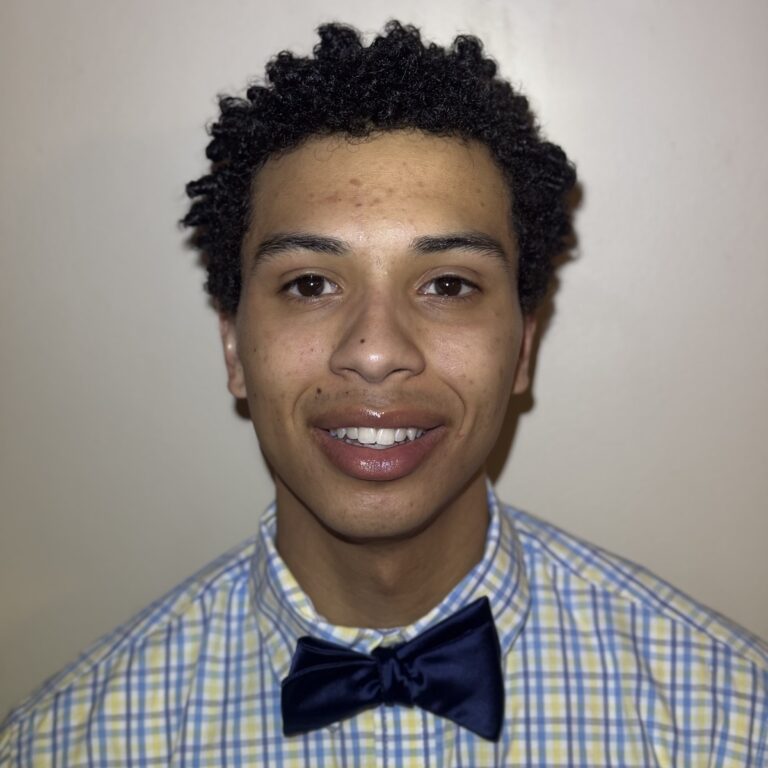 Photograph of a person wearing a blue and yellow plaid shirt with a dark blue bow tie, standing against a plain light-colored background.