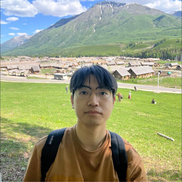 Photograph of a person standing on a green hillside with a village of wooden buildings behind them and tall mountains in the background under a blue sky with scattered clouds.